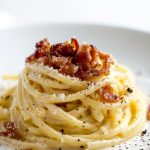 Spaghetti carbonara, sprinkled with fried guanciale, served on a white plate.