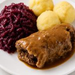 German rouladen covered in dark gravy with red cabbage and potato dumplings on a white plate.
