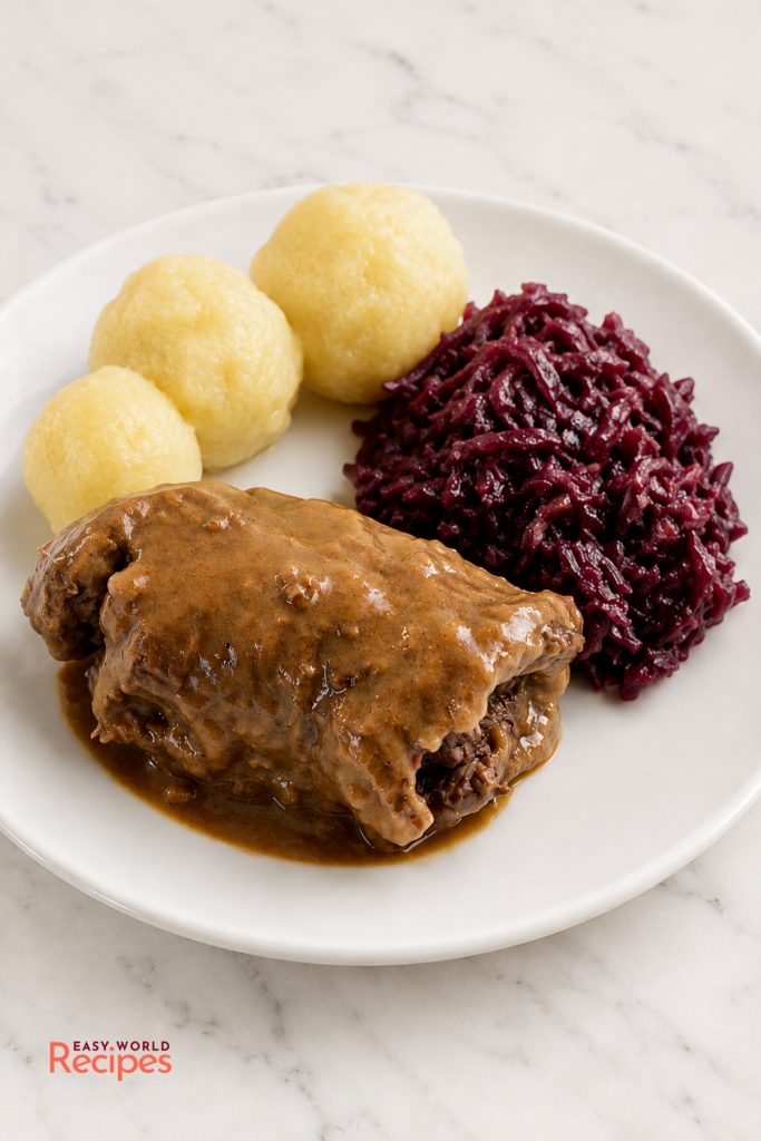 German rouladen sliced open to show the bacon, onion, and pickle filling with dark gravy, red cabbage, and potato dumplings.