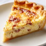 Slice of authentic French Quiche Lorraine with flaky pastry, creamy egg custard, and smoky bacon on a white plate.